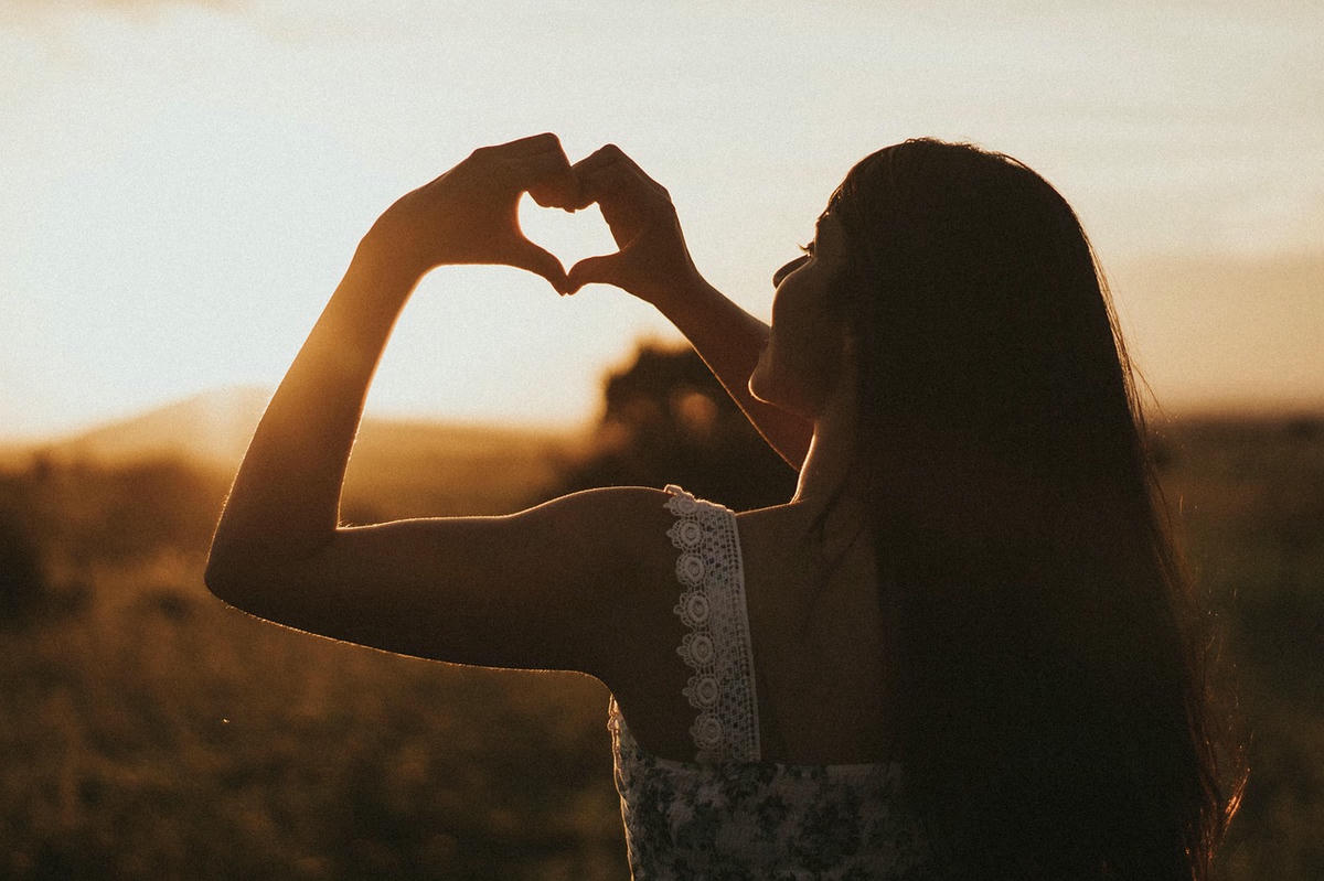 Vrouw die silhouet van een hart maakt met haar handen bij zonsondergang in een natuurlijke omgeving, symboliseert liefde en verbondenheid.