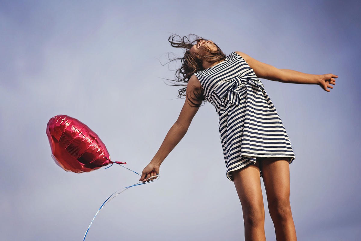 Vrouw in gestreepte jurk springt vrij met een hartvormige ballon tegen blauwe lucht – symbool voor vrijheid en liefde
